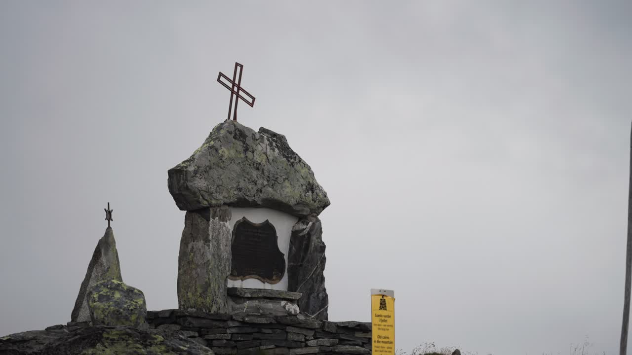 금속 크로스를 가진 기념비가 바위 위에 서 있으며, 산의 구름이 가득한 하늘을 배경으로 설정되어 있습니다.