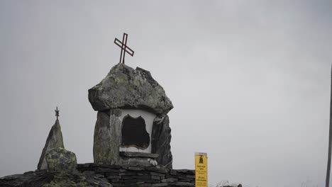 a monument with a metal crosse stands atop a rocky formation, set against a cloudy sky in the mountains