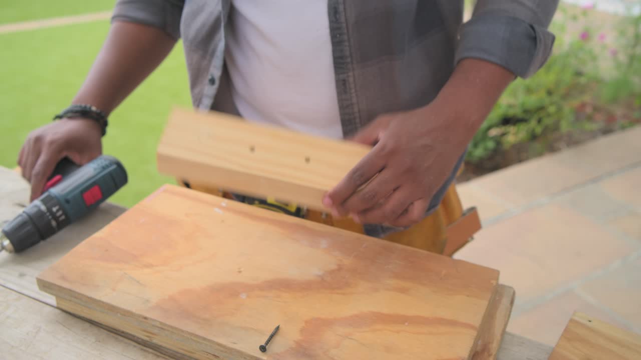Uomo con cintura degli attrezzi che usa un trapano, avvitando una vite, unendo pezzi di legno e ispezionando le parti sul patio.
