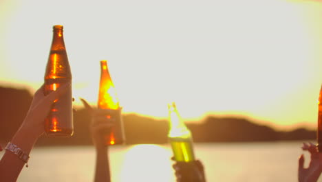 los jóvenes están bailando con las manos en alto y haciendo sonar la cerveza de las botellas de colores. esta es una fiesta loca al atardecer en la costa del río.