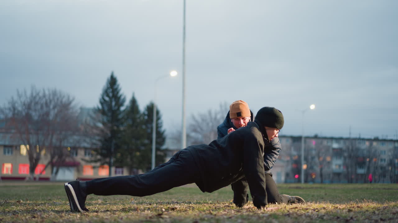 A coach closely supervises as a young boy dressed in black performs push-ups on a grassy field illuminated by outdoor light, the coach squats with one leg and gets up