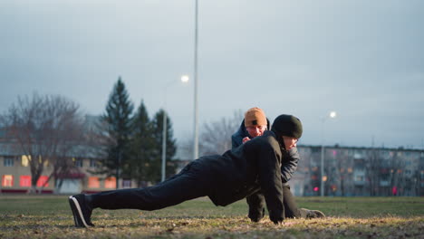 a coach closely supervises as a young boy dressed in black performs push-ups on a grassy field illuminated by outdoor light, the coach squats with one leg and gets up