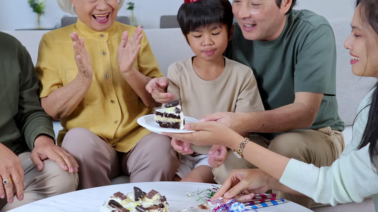 Three generations share birthday cake, wearing party hats, in a brightly lit living room