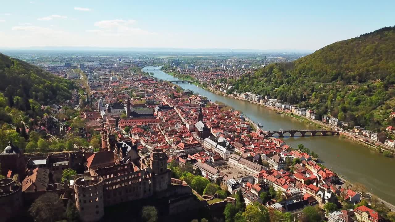 Aerial view capturing Heidelberg, Germany, featuring the castle, the Neckar river, Altstadt's housing red roofs, and a bridge amid lush green hills, drone orbiting shot