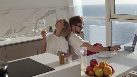 side view of bearded man working on laptop computer sitting at home on a kitchen with panoramic windows. young blonde wife sitting from behind and enjoying eating her apple in the morning