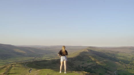 stabilised shot of young blonde woman jogging along the path on top of mam tor, castleton, peak district, england before stopping to admire the view of green rolling hills and blue skies