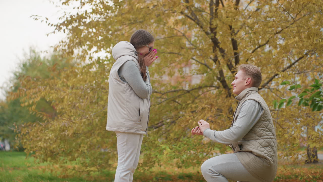 Lovers in matching beige outfits stand outdoors as man kneels down to propose with autumn leaves swaying in background woman surprised covers mouth
