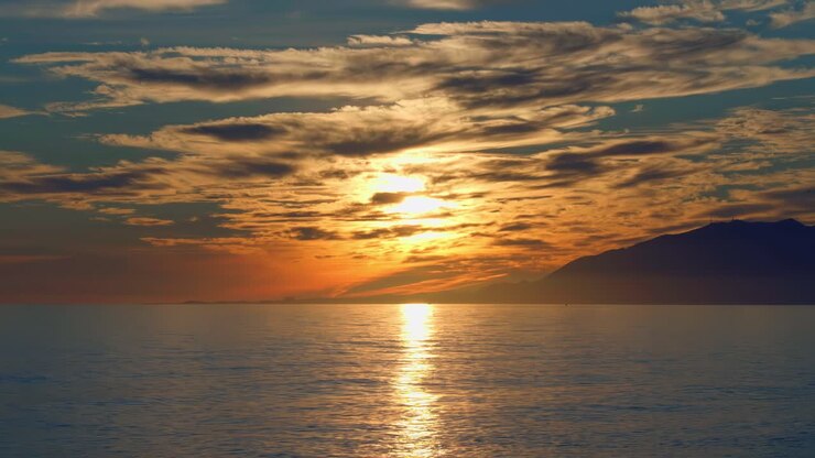 Dramatic sunset with calm ocean waves, clouds, and mountains on the background