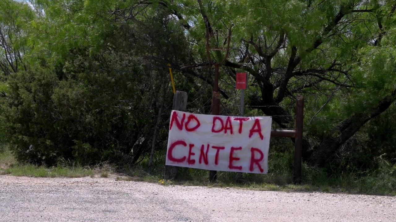 Un cartello bianco fatto in casa, dipinto audacemente con 'No Data Center' in rosso, si erge su uno sfondo di alberi verdi lussureggianti. Significa la resistenza della comunità locale e le preoccupazioni ambientali riguardo a proposte.