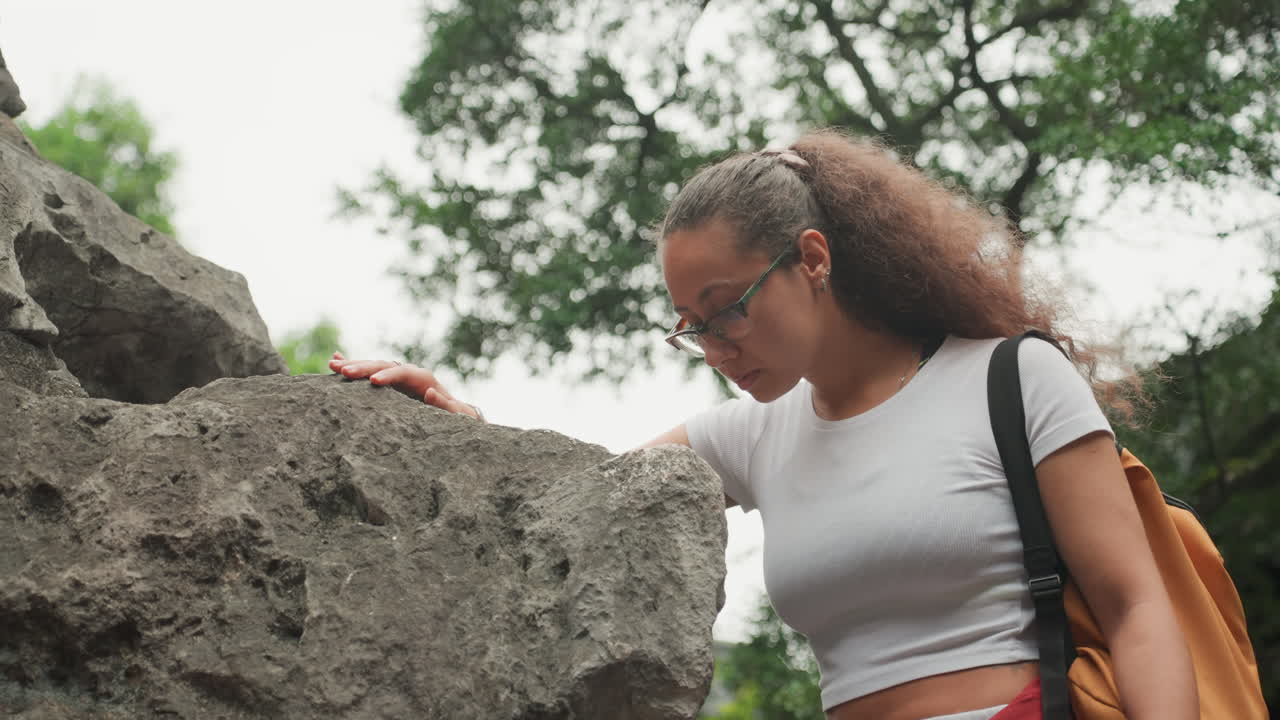 Young woman examines rough rock outdoors, backpack and eyeglasses visible, gentle hand tracing coarse stone texture, curly hair and casual outfit, focused student exploration and discovery in green