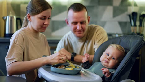 mother gives a spoon with pureed veggies to her baby. daddy sits behind looking at his lovely son.