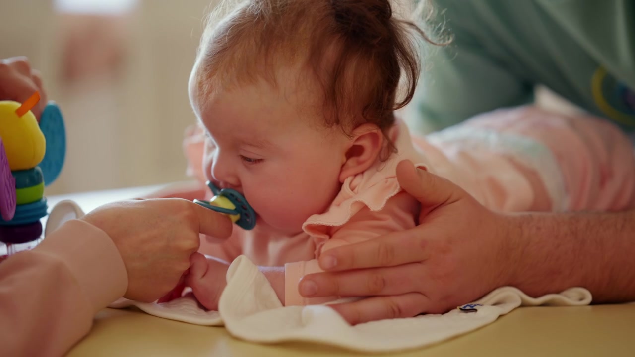 Um bebê deita-se confortavelmente enquanto observa um brinquedo colorido e faz exercícios de mobilidade com um cuidador.