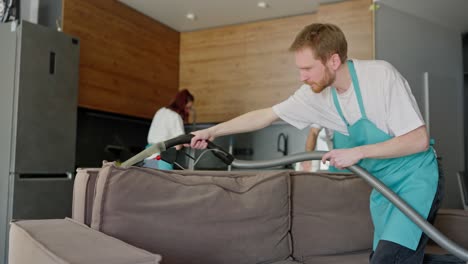 confident male blond cleaner in a white t-shirt and blue apron cleans a brown sofa using a vacuum cleaner with his colleagues in a modern studio apartment