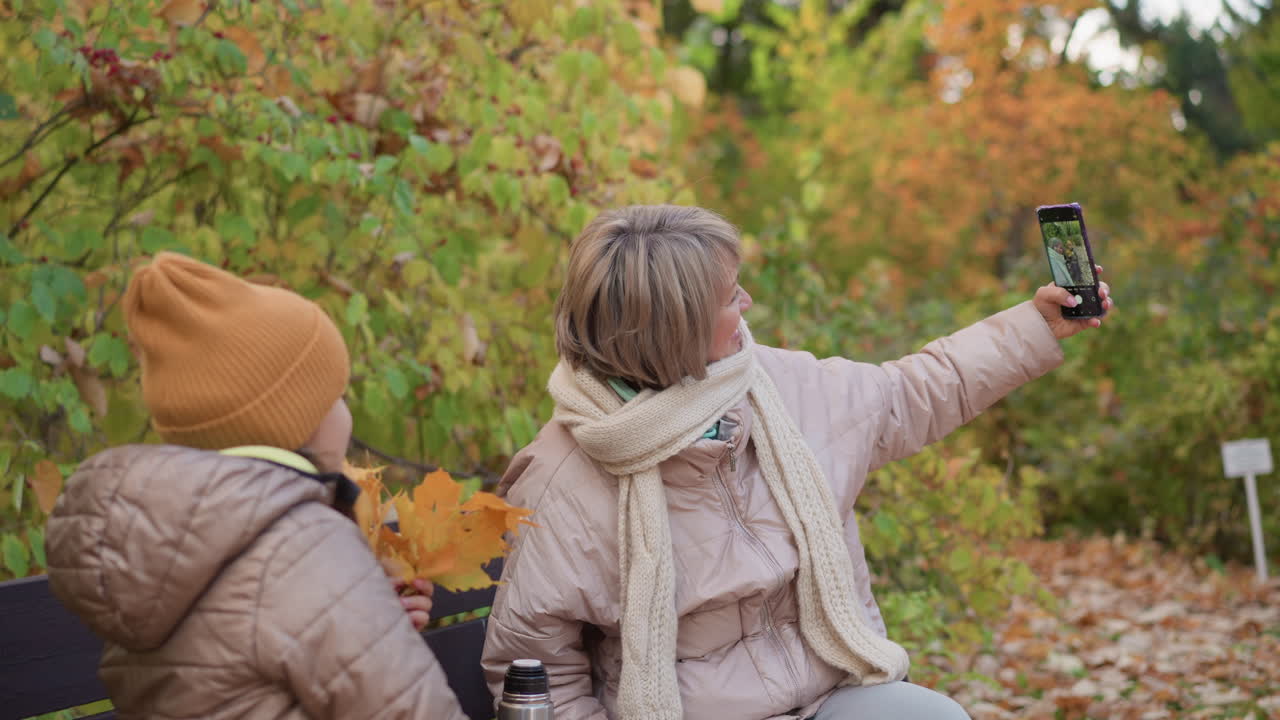 Mother in beige scarf and winter coat extends arm taking selfie with daughter holding maple leaves, both sitting close on wooden bench surrounded by golden autumn foliage in serene forest park
