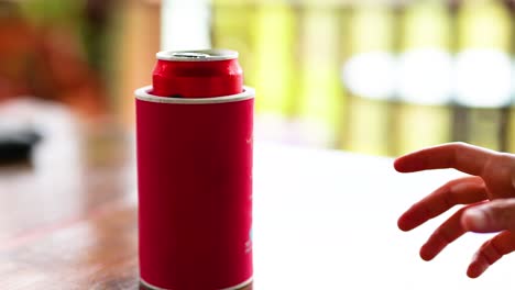 a hand reaches for a red can on a wooden table in a bright, blurred background, suggesting a relaxed atmosphere