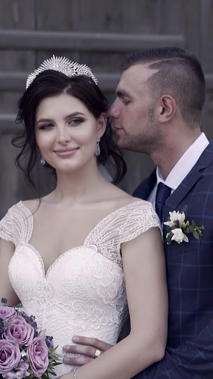 slow motion handsome fiance in suit hugs pretty bride in tiara with purple bouquet and whispers in ear