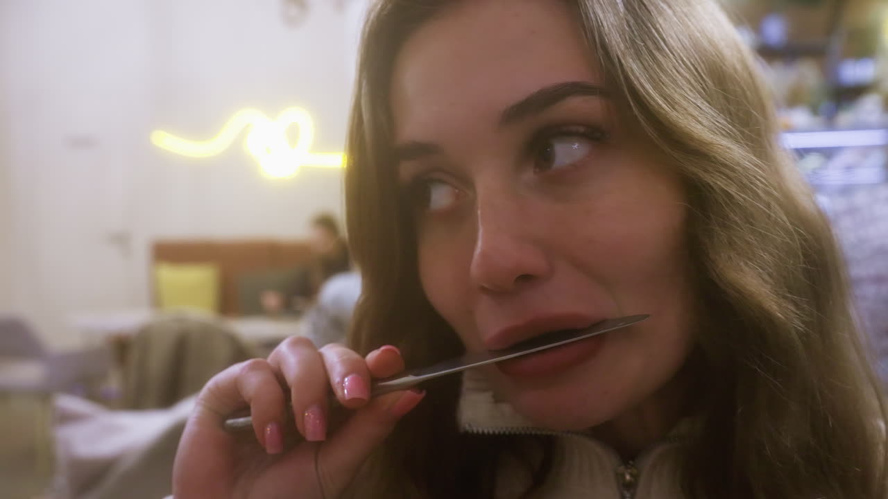Close-up of woman in beige shirt at cafe licking small knife with a playful or relaxed expression. Soft lighting and cozy ambiance in background, capturing intimate moment in cafe setting