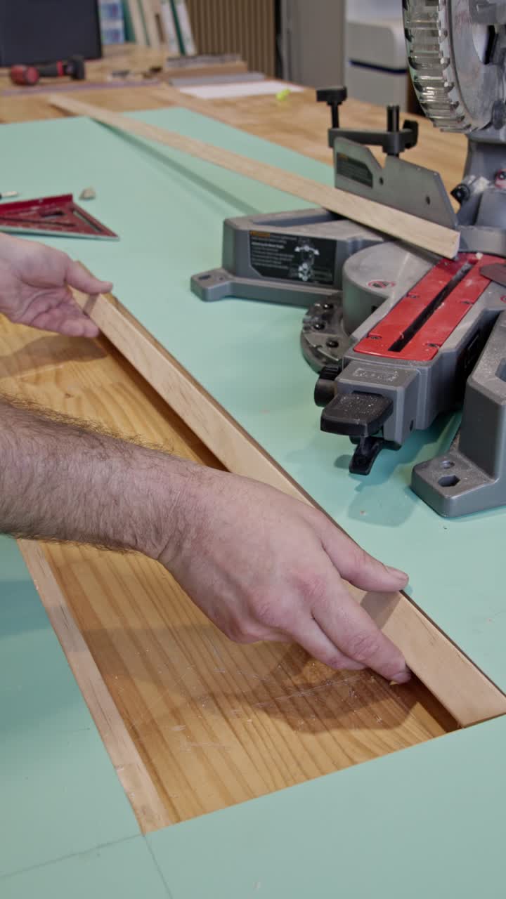 Vertical portrait shot of a carpenter aligning a wooden door trim piece on a workbench beside a miter saw. Hands carefully position the timber along the edge of a flat door panel
