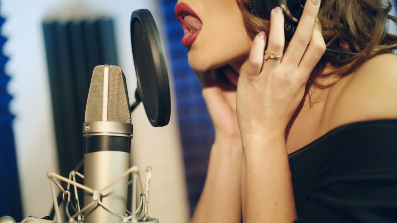 Beautiful woman with headphones singing close to the mic in the studio. Professional white and black microphone and female's face with bright lips. Close-up.