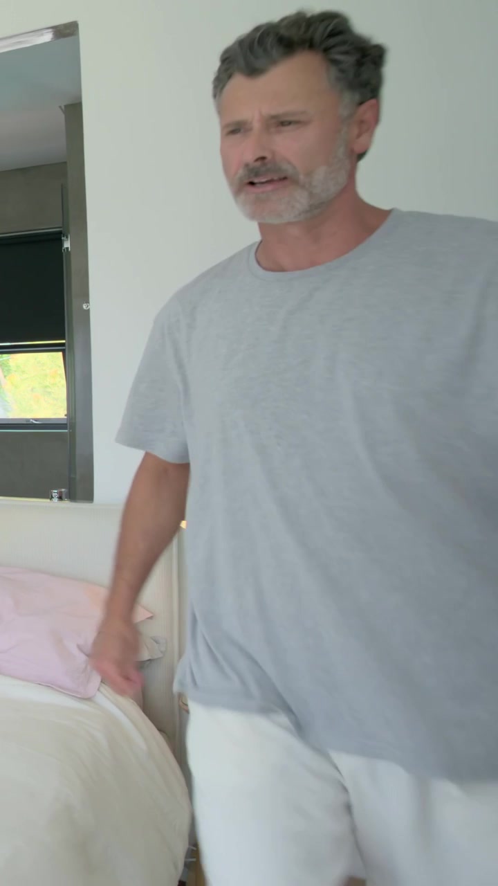 Video verticale: Un uomo in maglietta grigia entra da destra e rifà il letto con cuscini rosa, vicino al bagno, con spazio per il testo.