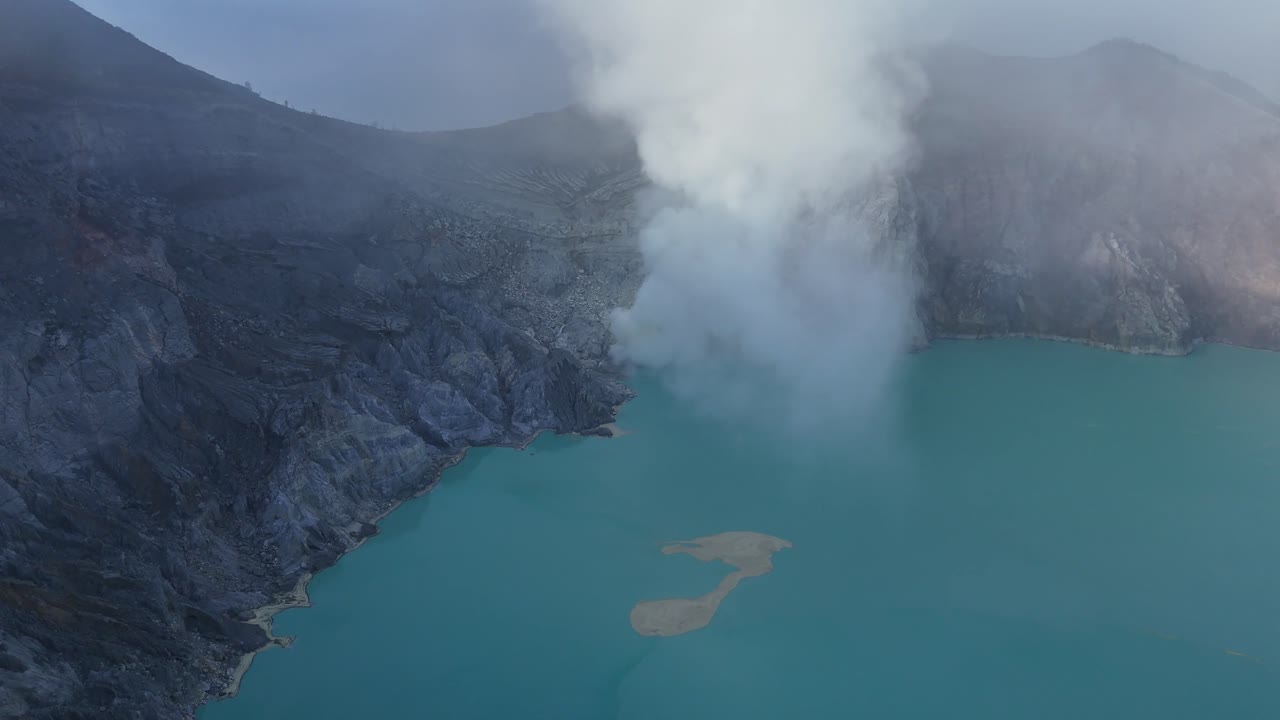 Drone view of Kawah Ijen crater lake in Java Indonesia, with turquoise waters and thick sulfur smoke rising from volcanic vents amid rugged terrain