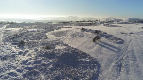 Snow-covered-rural-winter-countryside-track-footprint-shadows-terrain-aerial-view-slowly-lowering