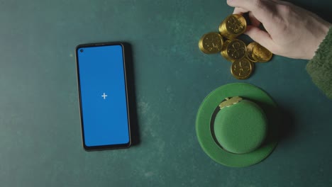 overhead studio shot of green leprechaun top hat with gold coins and blue screen mobile phone to celebrate st patricks day 2