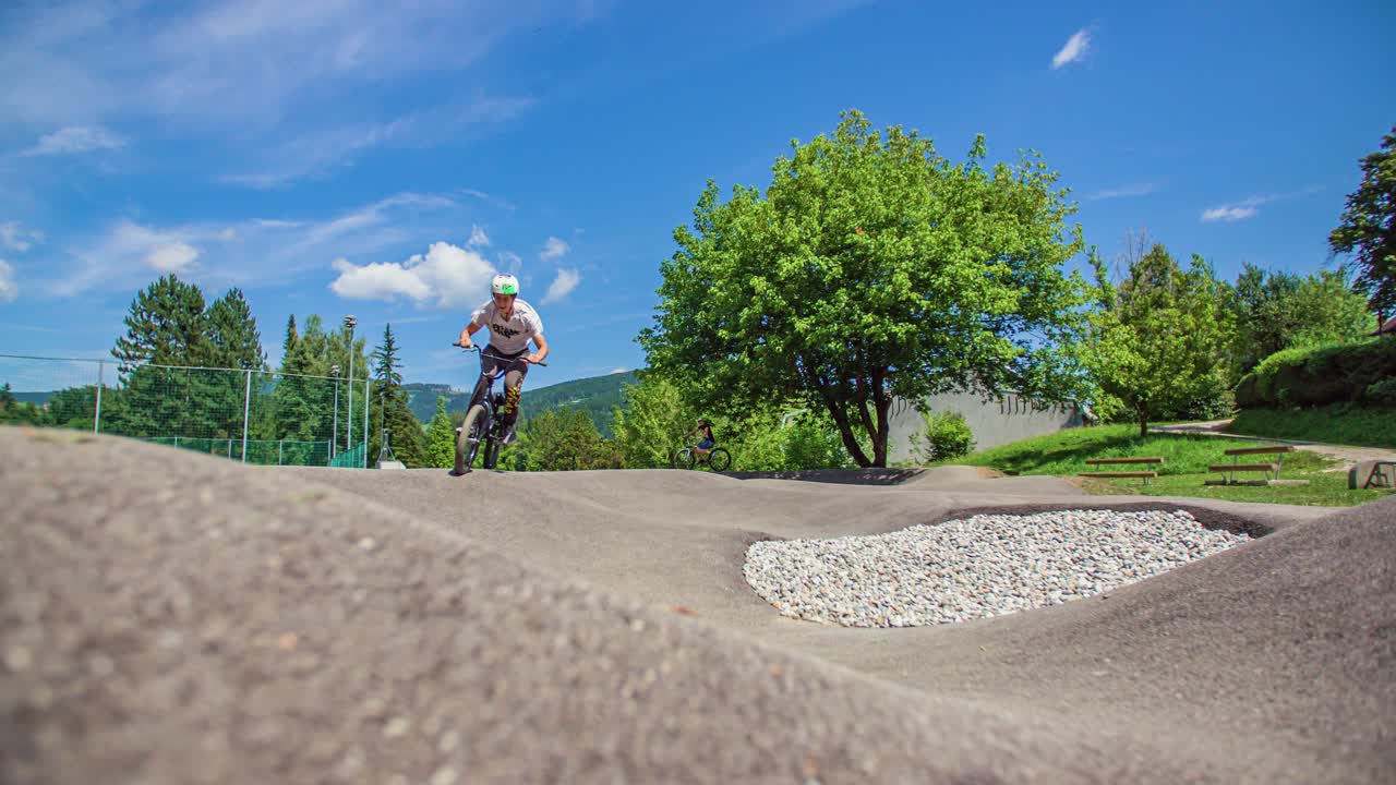 Un ciclista su una pump track si dirige verso la telecamera, salta e gira a destra superando l'inquadratura, al rallentatore, primo piano