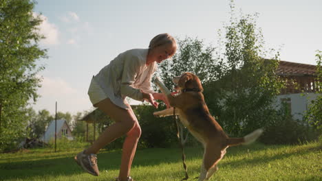 pet lover dancing playfully interacts with her dog in lush green garden under sunny weather, trying to kiss dog while it looks up happily, with buildings and trees in background