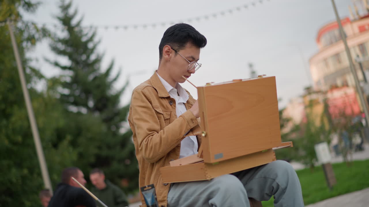 seorang ilustrator Asia melukis di taman kota dengan kotak kayu sebagai easel, fokus pada komposisi di bawahString Lights, pohon cemara dan bangunan pastel di latar belakang, orang-orang tampak buram di bangku, santai