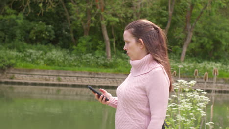 Woman-Outdoors-Walking-Along-Bank-Of-River-Looking-At-Mobile-Phone