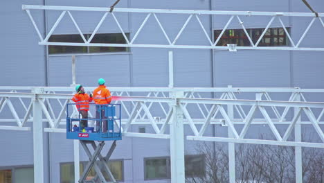 two workers dressed in orange overalls, on a crane in a building under construction