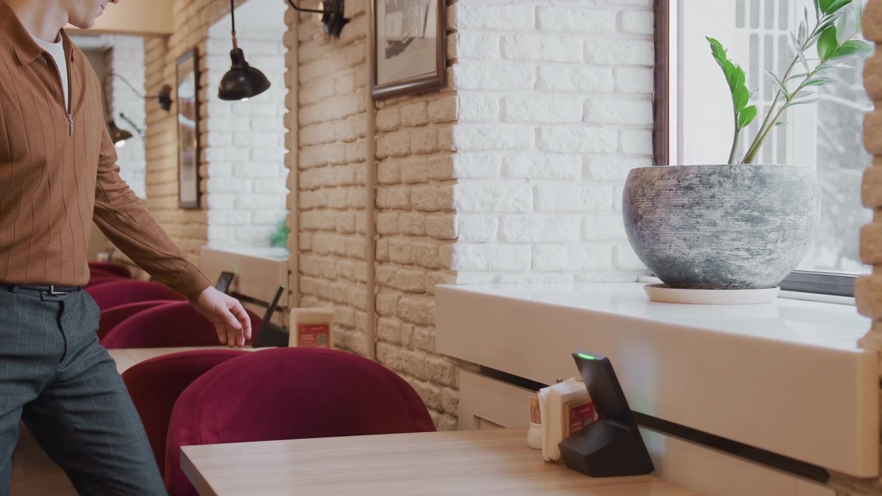 Un uomo con una camicia marrone si siede a un tavolo, poi si alza e si sposta per spingere indietro la sedia. L'ambientazione è un caffè interno con pareti di mattoni bianchi, sedie rosse, lampade a sospensione, quadri incorniciati sulla parete e luce naturale.