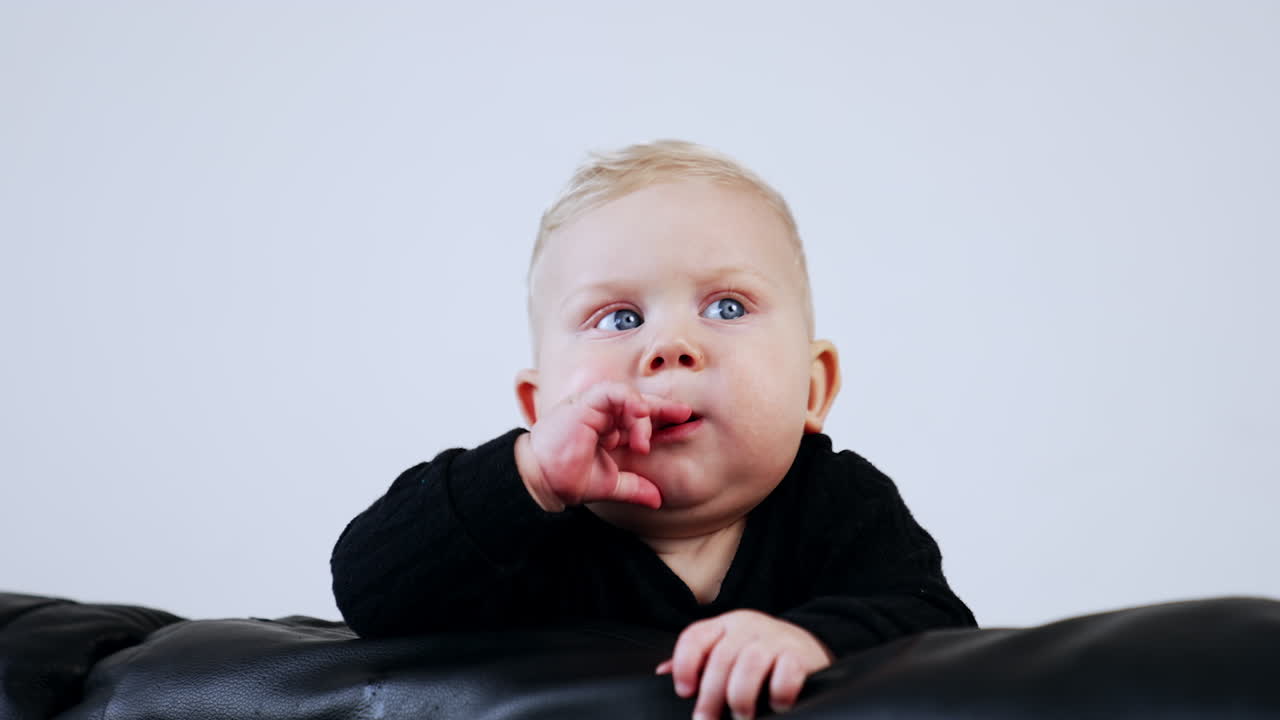 Adorable Caucasian baby with chubby face wearing black sweater. Little child on the black sofa at white backdrop.