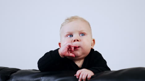 adorable caucasian baby with chubby face wearing black sweater. little child on the black sofa at white backdrop.