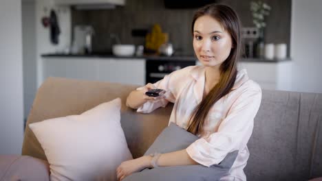 pretty brunette woman is resting on a couch in living room in holiday and switching tv channels by remote control