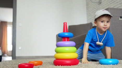 smiling caucasian baby boy comes up to a toy pyramid. kid in a cap sits down on the floor to play with his toy. low angle view.