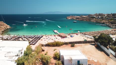 aerial drone orbit shot with super paradise beach in mykonos, greece during a clean sky sunny day with tourists and water sports residents on the hill