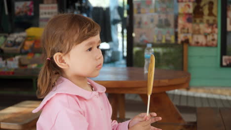 Cute-Girl-Eats-and-Licks-Dalgona-Sugar-Candy-on-Stick-with-Heart-Shape-at-Gyeongamdong-Railroad-Town-Old-Market---Portrait-Slow-motion