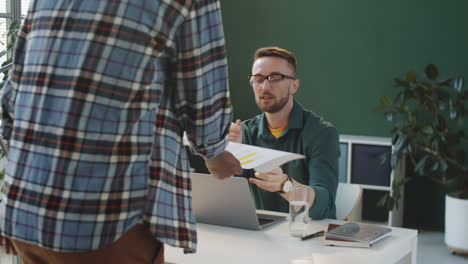 Compañeros-De-Trabajo-Masculinos-Dándose-La-Mano-Y-Discutiendo-Papel-En-La-Oficina