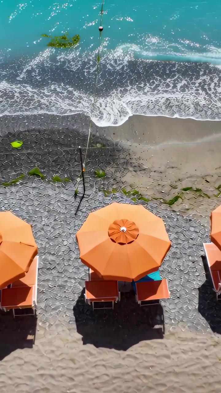 Video verticale: La telecamera si sposta verso una boa, onde che creano schiuma vicino a ombrelloni e lettini arancioni