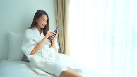 Woman-relaxing-on-bed-using-phone,-smiling-as-she-scrolls-seated-near-window-with-morning-light