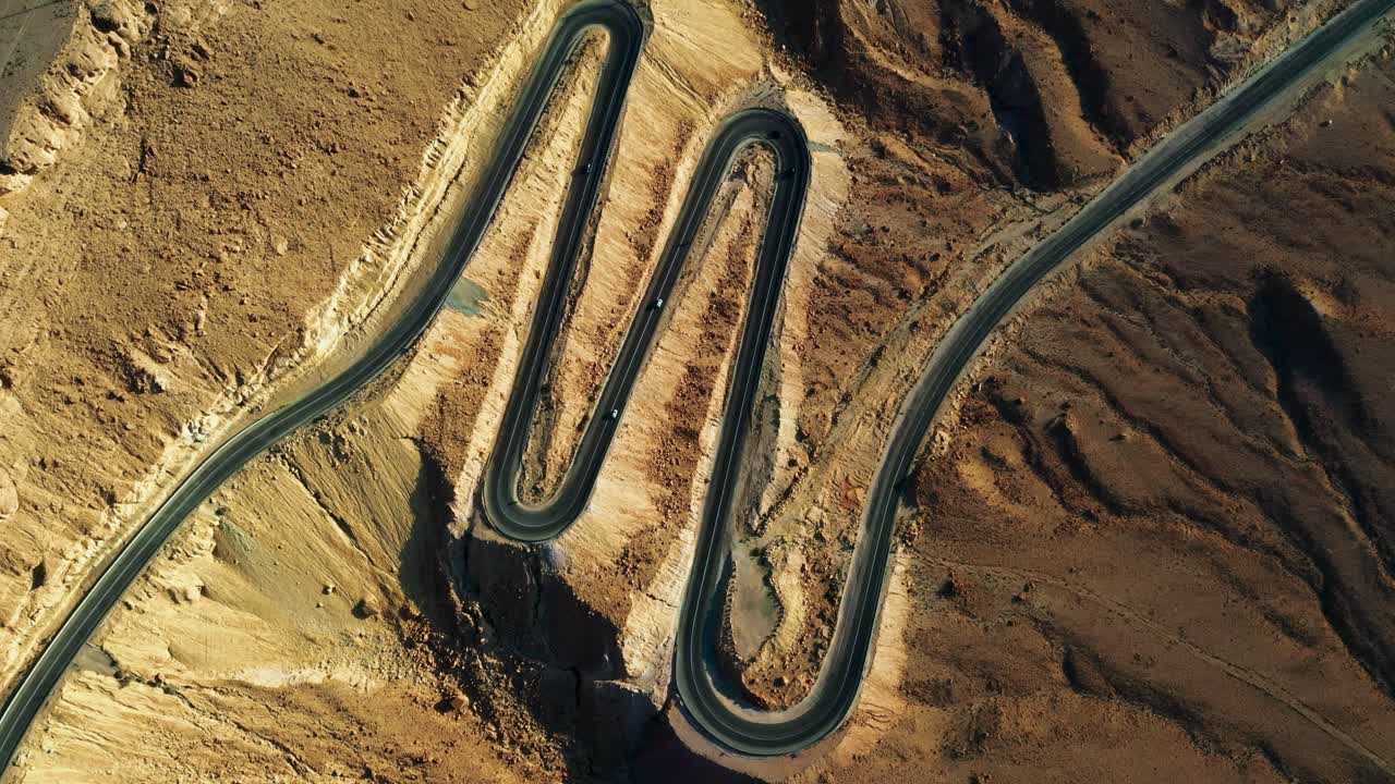 5대의 차량이 이스라엘의 모래색 미츠페 라몬의 산간 풍경으로 내리막길을 굽이치는 날카로운 머리핀 사이의 검은 아스팔트를 주행합니다.