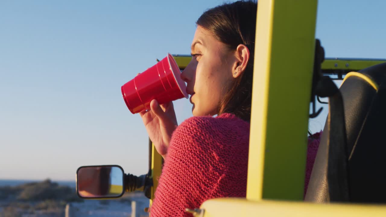 mujer caucásica feliz sentada en un buggy de playa junto al mar bebiendo