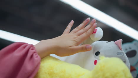 lady in pink gently touches a white stuffed toy's hand on a shelf in a brightly lit store, she touch the soft fabric carefully, with other toys around in the store, with the store well lit up