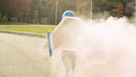 vista posteriore di un ragazzino che indossa ali di aeroplano di cartone con fumo artificiale dietro che corre nel parco e gioca come pilota 1