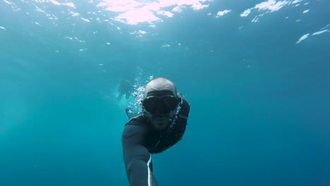 White-male-freediver-with-beard-does-360-degree-barrel-roll-while-holding-a-selfie-stick-in-blue-tropical-waters-with-sunlight-behind-him