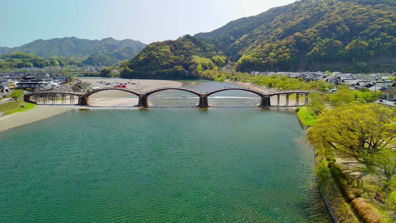 Aerial pull-back reveals the iconic Kintai Bridge in Iwakuni, Japan. The historic five-arched wooden structure spans the Nishiki River, set against a backdrop of lush mountains on a sunny day
