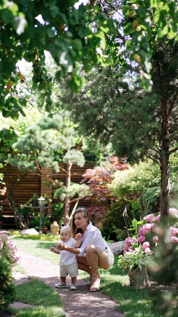 Mother is walking with her little baby son in the garden. Mom is leading her child by the hand and points at camera. Vertical video.