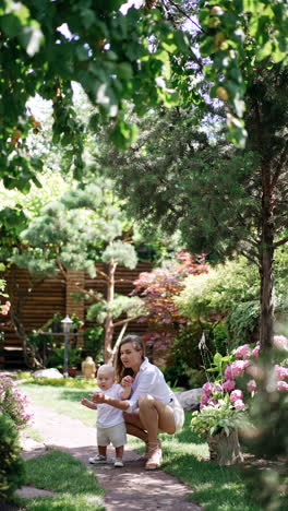 mother is walking with her little baby son in the garden. mom is leading her child by the hand and points at camera. vertical video.